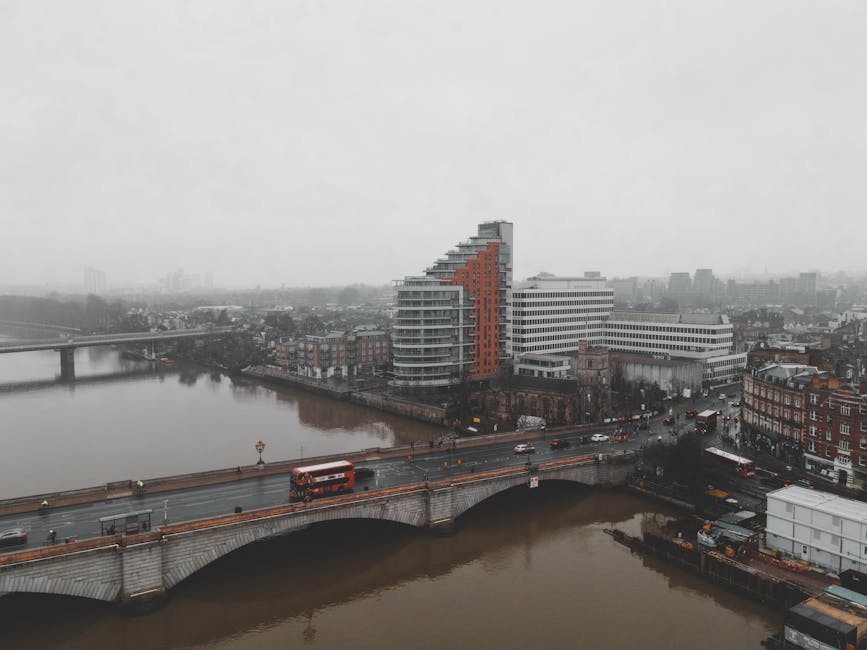 A red double-decker bus with route number 9 displaying 'Hammersmith' on its electronic sign is traveling along a city street. The bus is heading towards Hammersmith Bridge, with a mix of pedestrians w