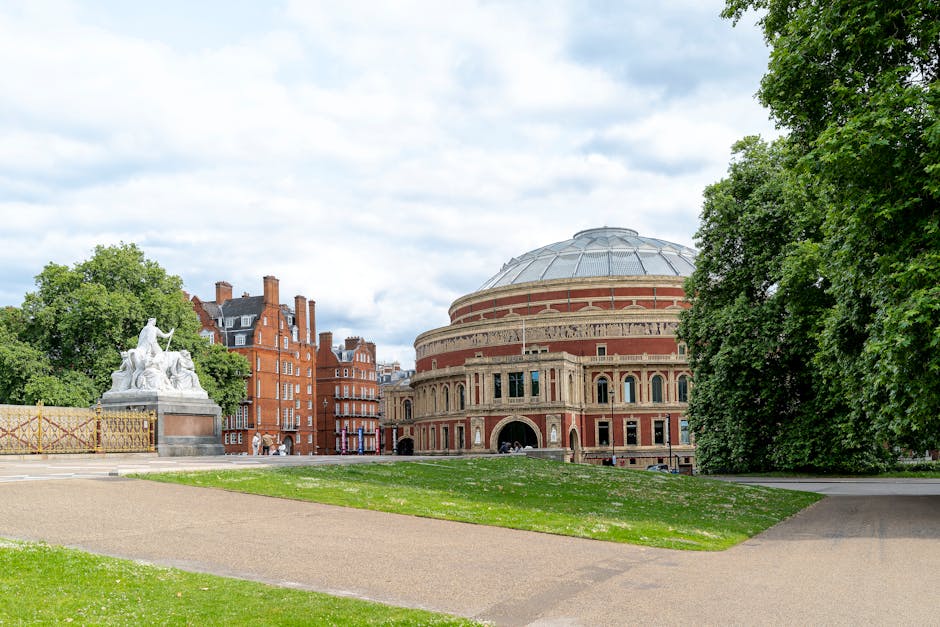 A spacious outdoor park scene with large mature trees featuring broad green foliage providing shade over the open ground. In the background, there is a small wooden bandstand with a canopy roof, surro