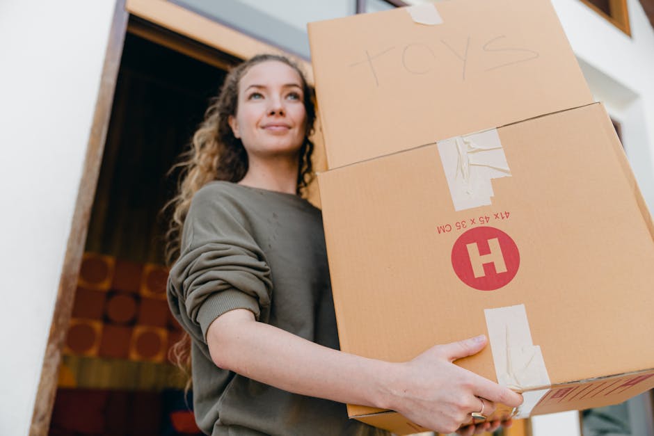 A man and a woman standing inside a residential building next to a doorway, both holding cardboard boxes loaded with household items. The man, dressed in a maroon shirt and orange pants, presents a la
