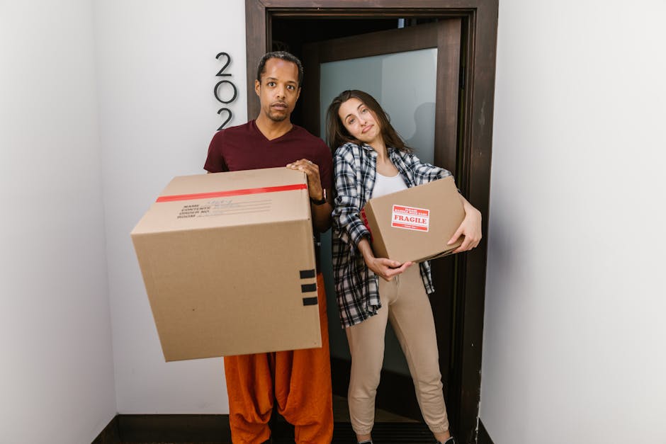 Two women are standing indoors amidst a collection of packed cardboard boxes, some sealed with red and black packing tape, suggesting a home relocation or move. One woman, wearing beige trousers, a wh